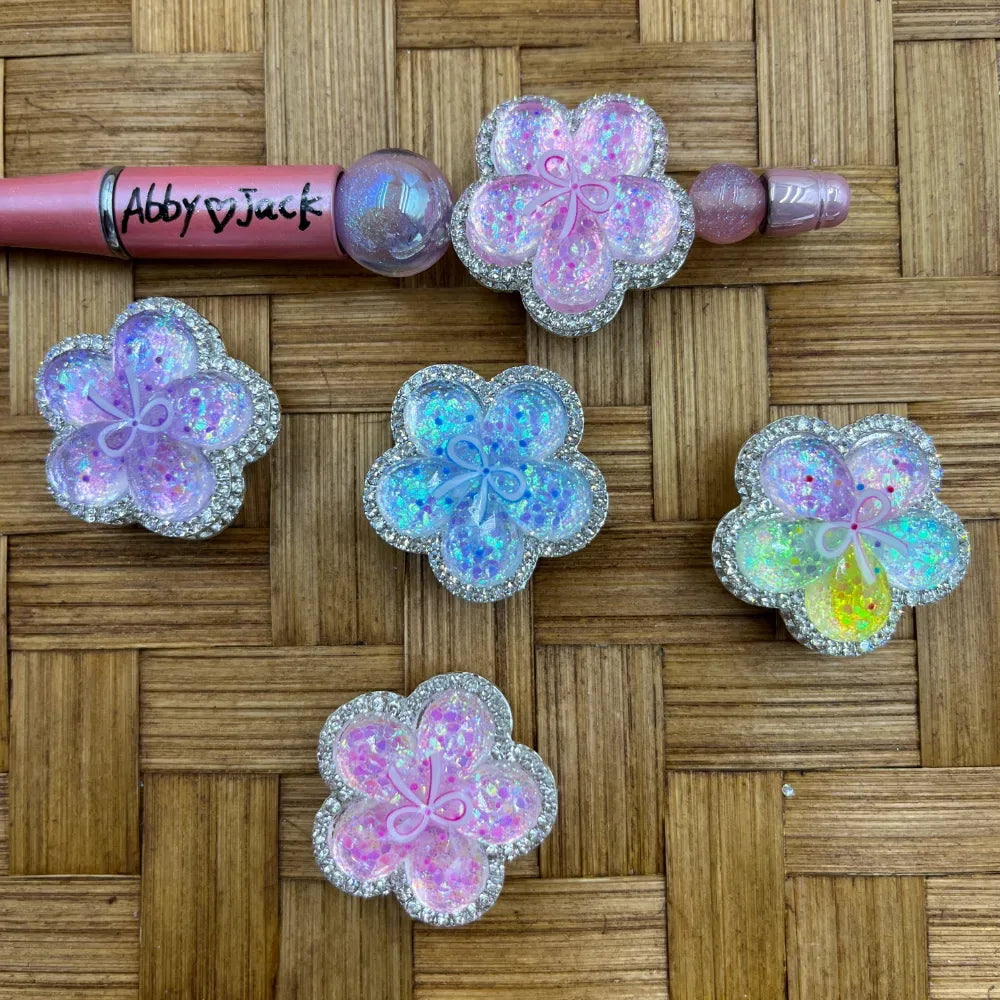 Five iridescent glitter flower-shaped resin flatback beads with silver rhinestone borders in pink, purple, blue, and rainbow colors, displayed on a wicker surface next to a beadable pen.