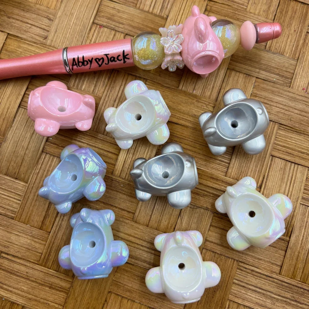 Assorted iridescent pearlescent bear shaped resin bead caps in pink, blue, silver, and white colors displayed on a woven surface next to a decorated beadable pen labeled Abby and Jack.