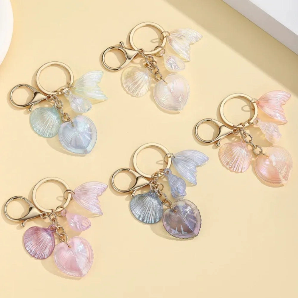 Five iridescent pastel resin keychain charms featuring heart-shaped shells, scallop shells, and mermaid tail pendants on gold lobster clasp key rings in blue, pink, and purple tones.