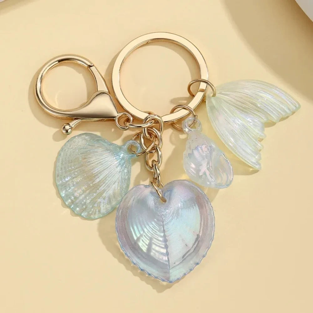 Five iridescent pastel resin keychain charms featuring heart-shaped shells, scallop shells, and mermaid tail pendants on gold lobster clasp key rings in blue, pink, and purple tones. - 4