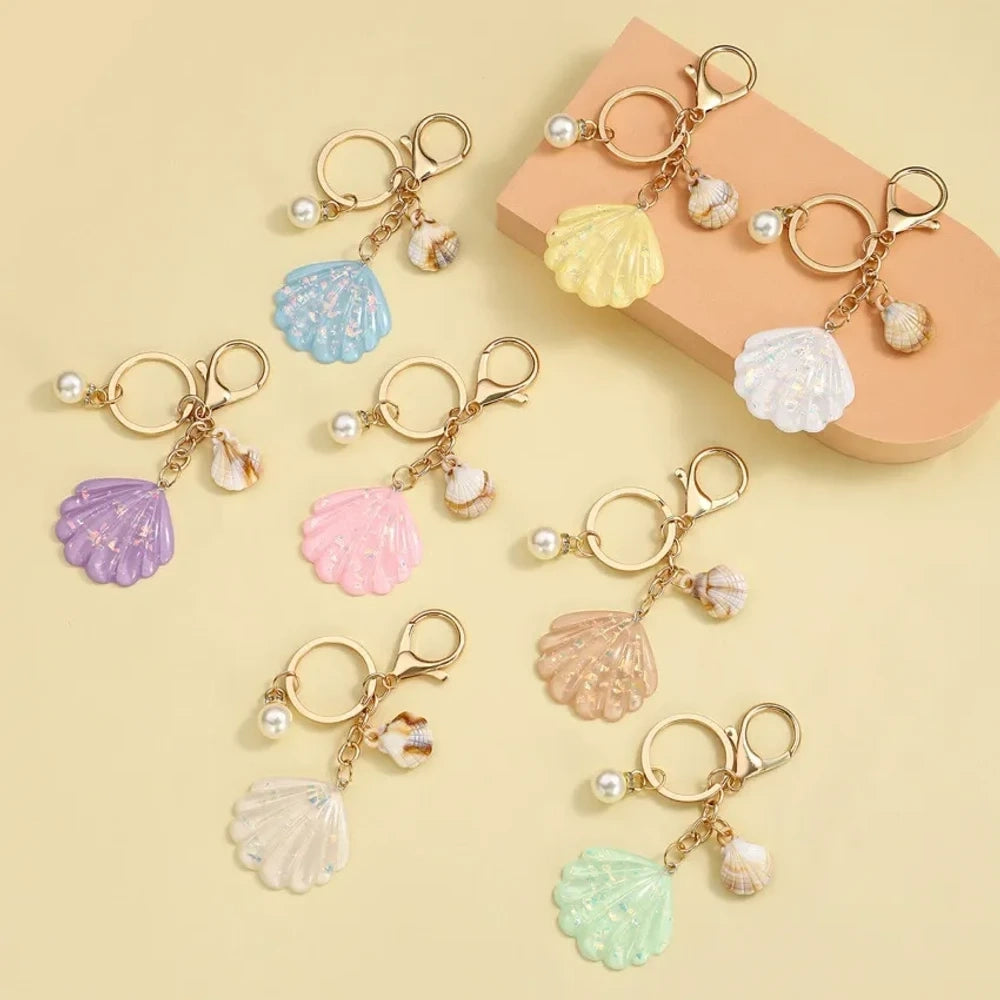 Collection of pastel iridescent resin scallop shell keychains in purple, pink, blue, yellow, white, mint, and blush tones with pearl beads and mini natural shell accents on gold-tone hardware, displayed on a cream background.