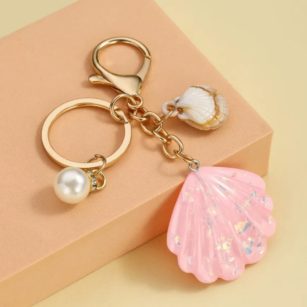 Collection of pastel iridescent resin scallop shell keychains in purple, pink, blue, yellow, white, mint, and blush tones with pearl beads and mini natural shell accents on gold-tone hardware, displayed on a cream background. - 1