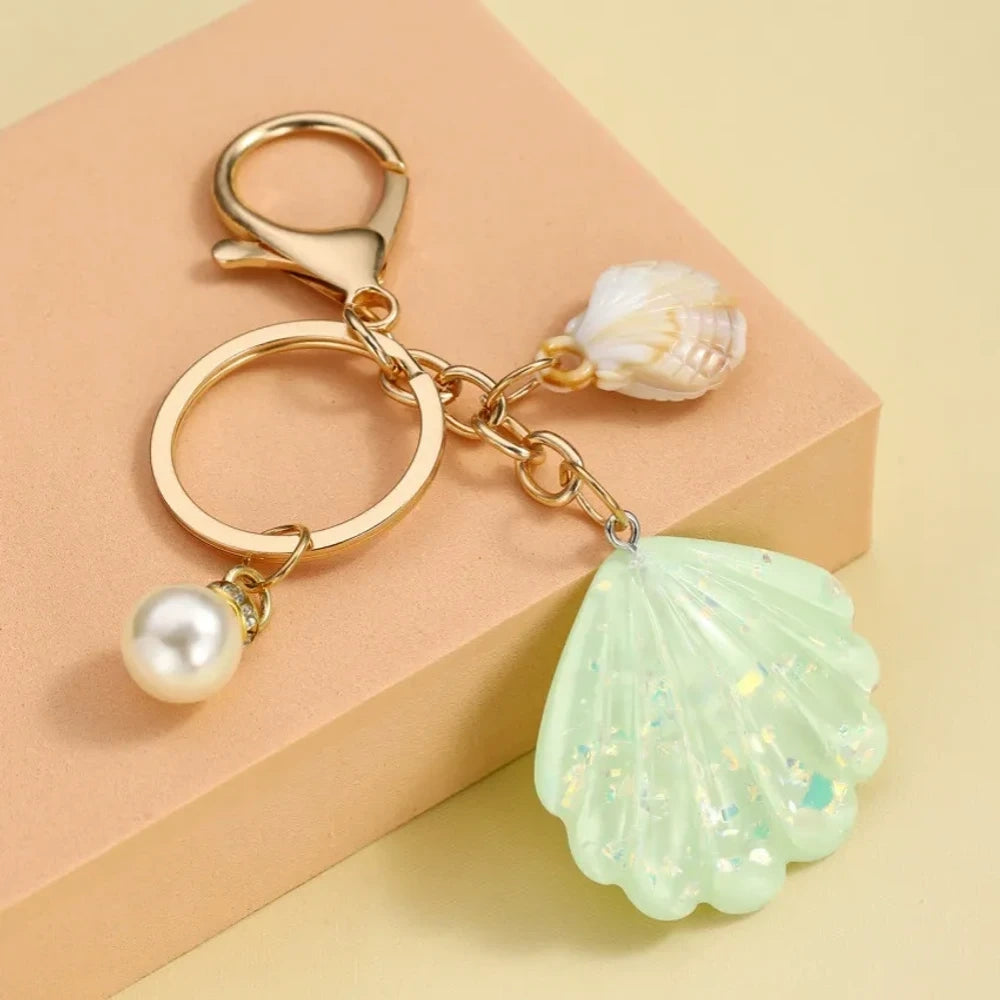 Collection of pastel iridescent resin scallop shell keychains in purple, pink, blue, yellow, white, mint, and blush tones with pearl beads and mini natural shell accents on gold-tone hardware, displayed on a cream background. - 2