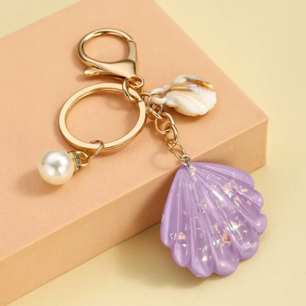 Collection of pastel iridescent resin scallop shell keychains in purple, pink, blue, yellow, white, mint, and blush tones with pearl beads and mini natural shell accents on gold-tone hardware, displayed on a cream background. - 3