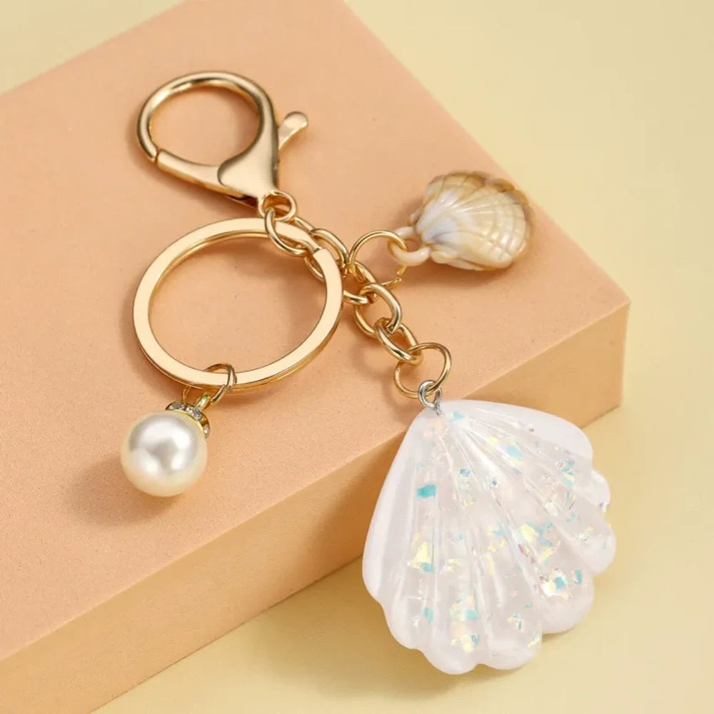 Collection of pastel iridescent resin scallop shell keychains in purple, pink, blue, yellow, white, mint, and blush tones with pearl beads and mini natural shell accents on gold-tone hardware, displayed on a cream background. - 4