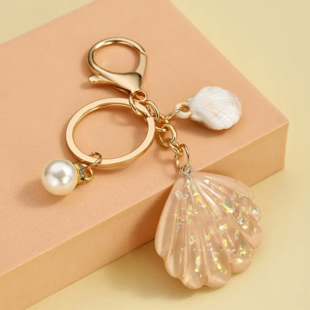 Collection of pastel iridescent resin scallop shell keychains in purple, pink, blue, yellow, white, mint, and blush tones with pearl beads and mini natural shell accents on gold-tone hardware, displayed on a cream background. - 5