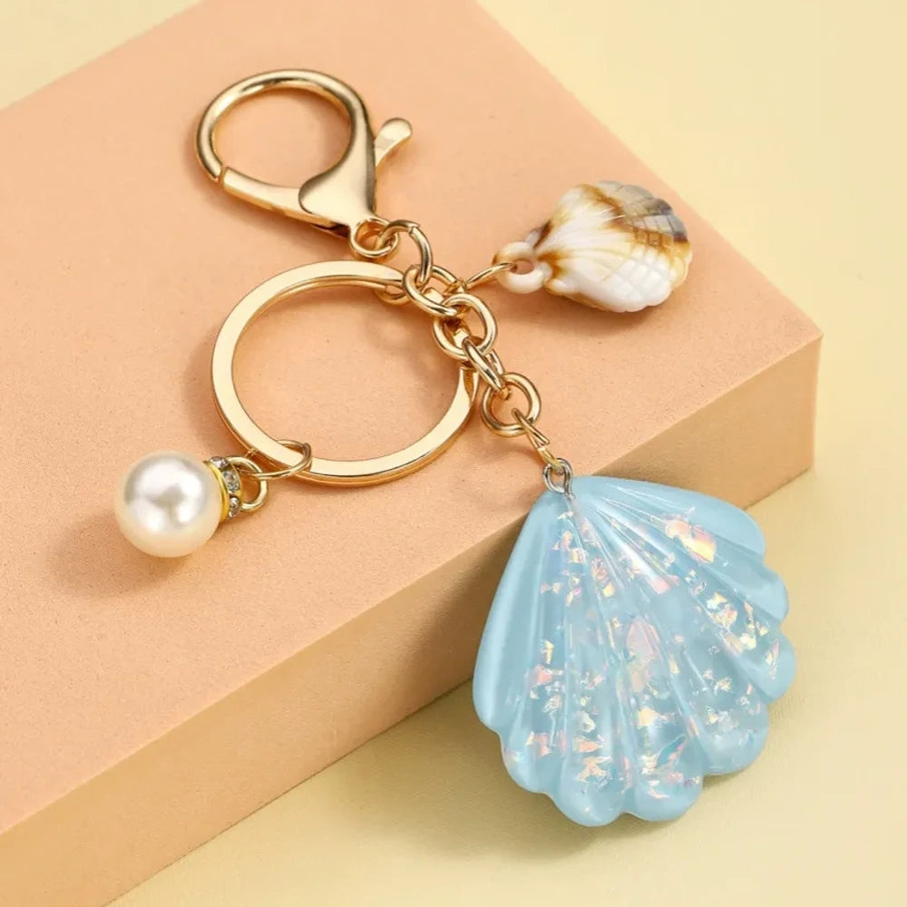 Collection of pastel iridescent resin scallop shell keychains in purple, pink, blue, yellow, white, mint, and blush tones with pearl beads and mini natural shell accents on gold-tone hardware, displayed on a cream background. - 6