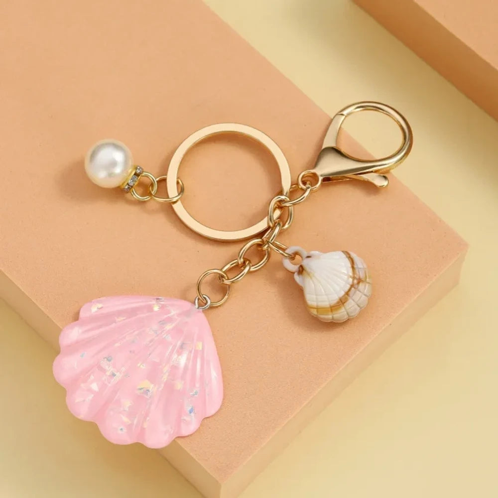 Collection of pastel iridescent resin scallop shell keychains in purple, pink, blue, yellow, white, mint, and blush tones with pearl beads and mini natural shell accents on gold-tone hardware, displayed on a cream background. - 7