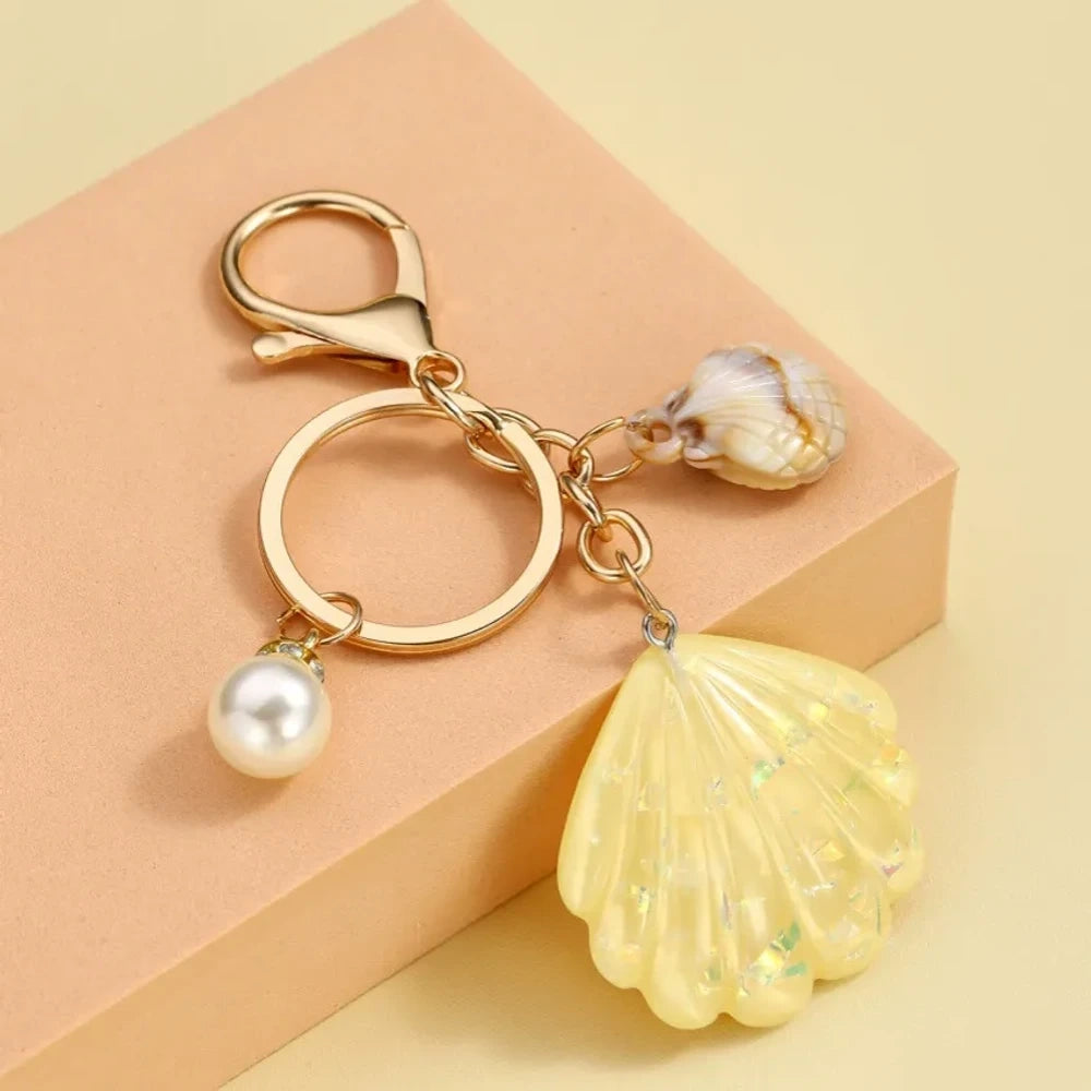Collection of pastel iridescent resin scallop shell keychains in purple, pink, blue, yellow, white, mint, and blush tones with pearl beads and mini natural shell accents on gold-tone hardware, displayed on a cream background. - 8
