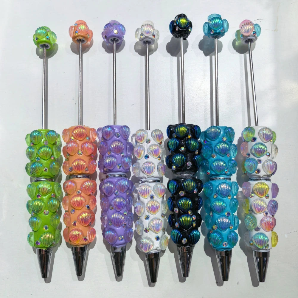 Seven iridescent seashell clay beadable pens in green, orange, purple, white, black, teal, and clear with AB rhinestone accents and silver metal tips arranged side by side on a white background.
