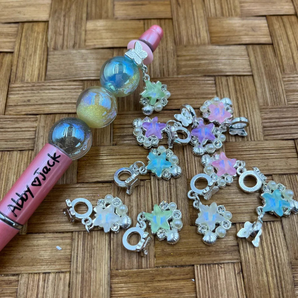 Iridescent opal star flower silver bail beads with butterfly charms and pearl accents displayed next to a beadable glitter ball pen on a wicker surface