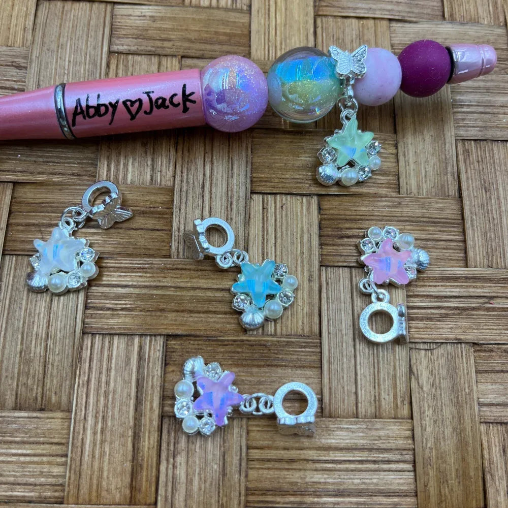 Iridescent opal star flower silver bail beads with rhinestones and pearl accents in white, blue, and pink, displayed next to a beadable pen on a woven bamboo surface.