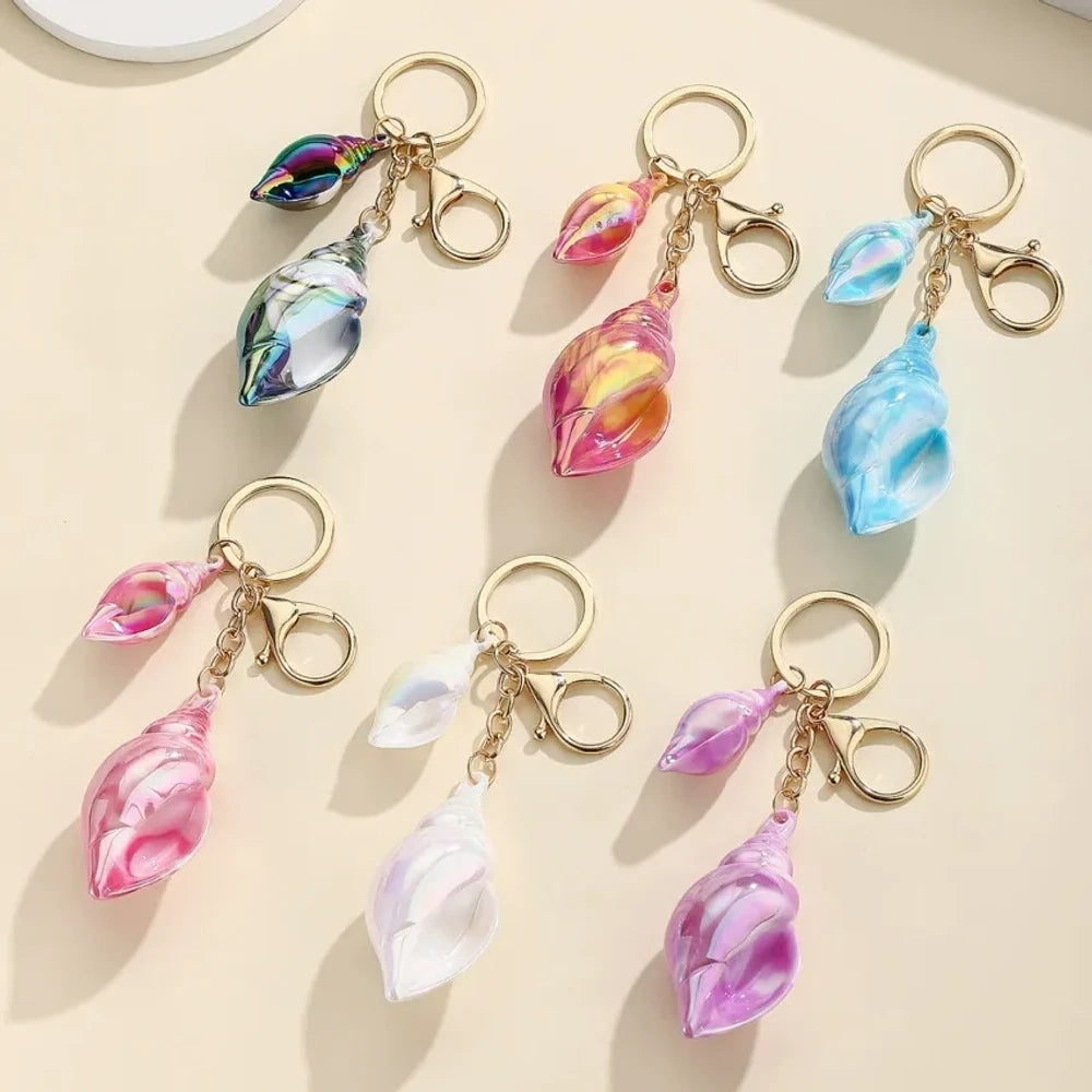Six colorful iridescent swirl resin conch shell keychain charms in rainbow, pink, blue, white, and purple with gold lobster clasp hardware on a cream background.