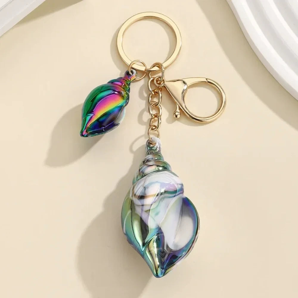 Six colorful iridescent swirl resin conch shell keychain charms in rainbow, pink, blue, white, and purple with gold lobster clasp hardware on a cream background. - Black