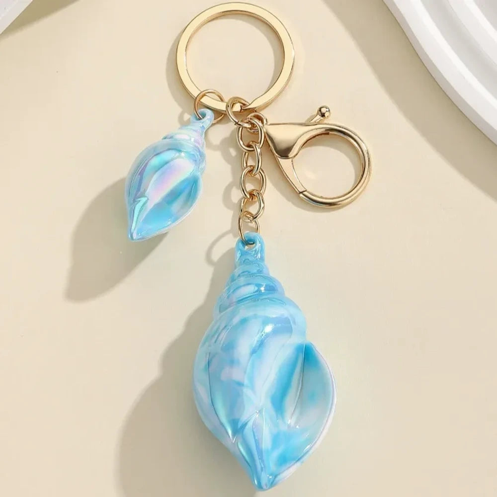 Six colorful iridescent swirl resin conch shell keychain charms in rainbow, pink, blue, white, and purple with gold lobster clasp hardware on a cream background. - Blue