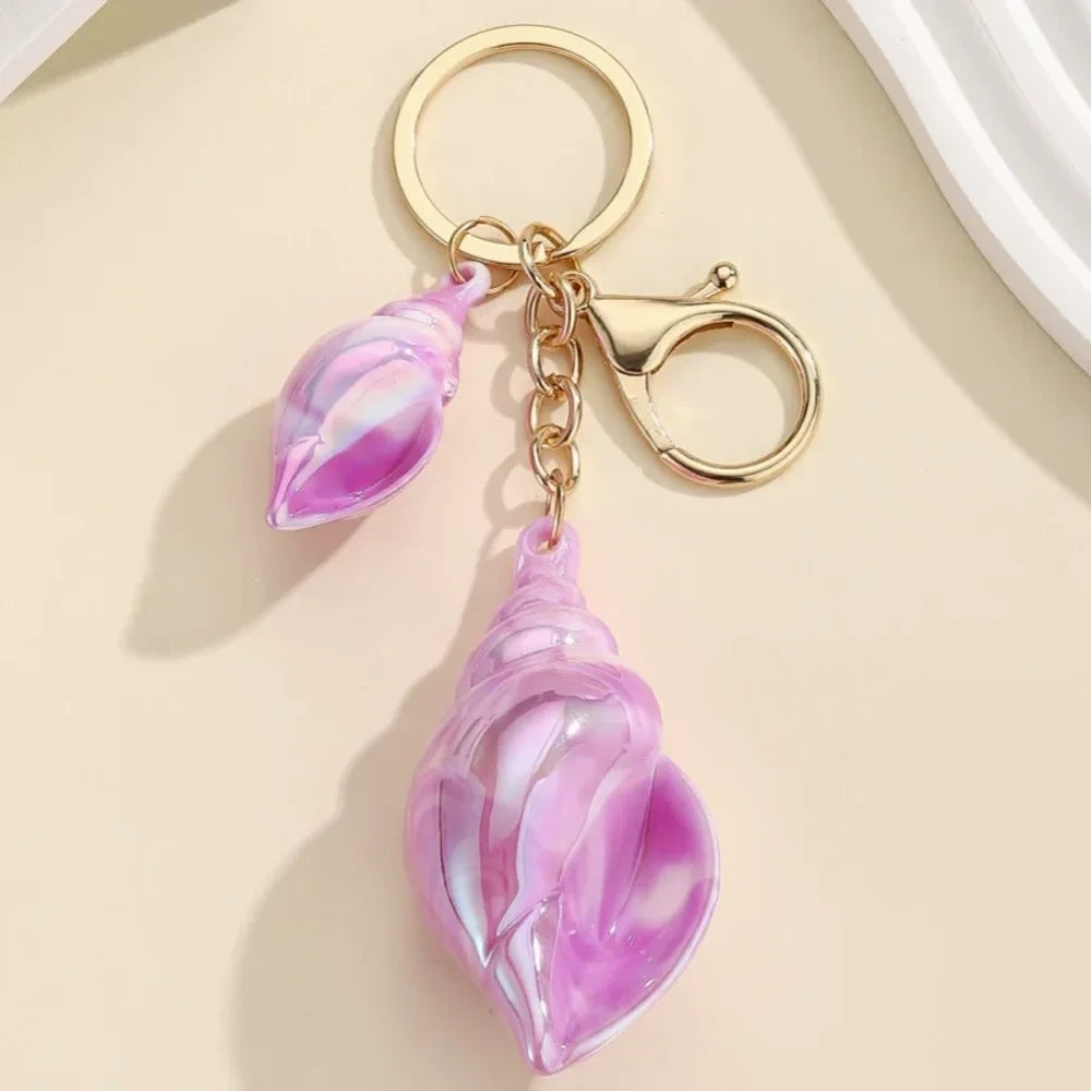 Six colorful iridescent swirl resin conch shell keychain charms in rainbow, pink, blue, white, and purple with gold lobster clasp hardware on a cream background. - Purple