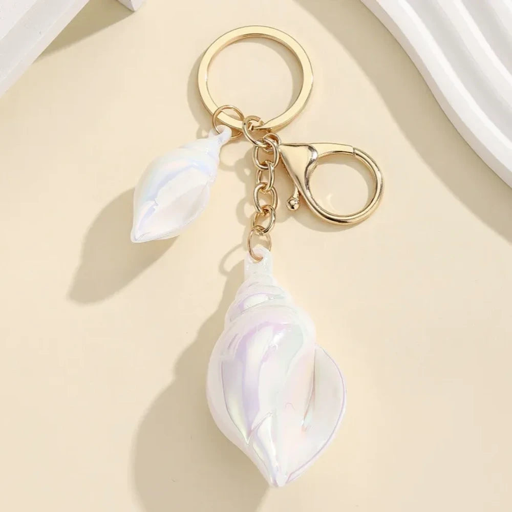 Six colorful iridescent swirl resin conch shell keychain charms in rainbow, pink, blue, white, and purple with gold lobster clasp hardware on a cream background. - White