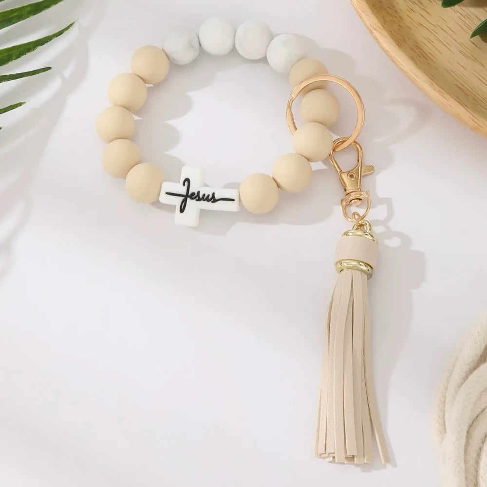 Four Jesus cross silicone bead bracelet keychains with leather tassels in beige, pink, blue, and gray marble color combinations on a white surface. - 1