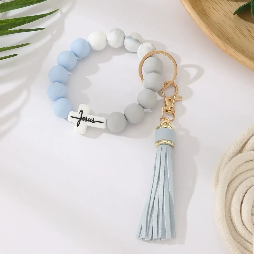 Four Jesus cross silicone bead bracelet keychains with leather tassels in beige, pink, blue, and gray marble color combinations on a white surface. - 2