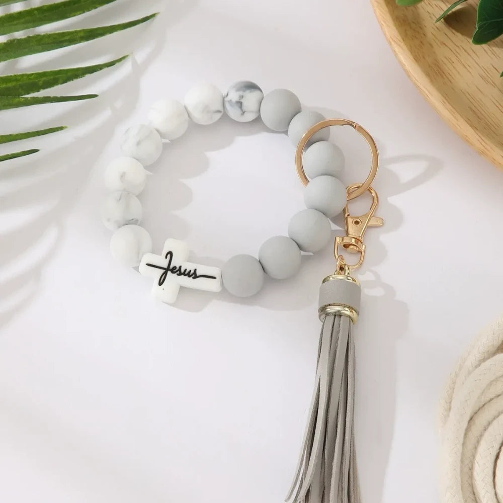 Four Jesus cross silicone bead bracelet keychains with leather tassels in beige, pink, blue, and gray marble color combinations on a white surface. - 3