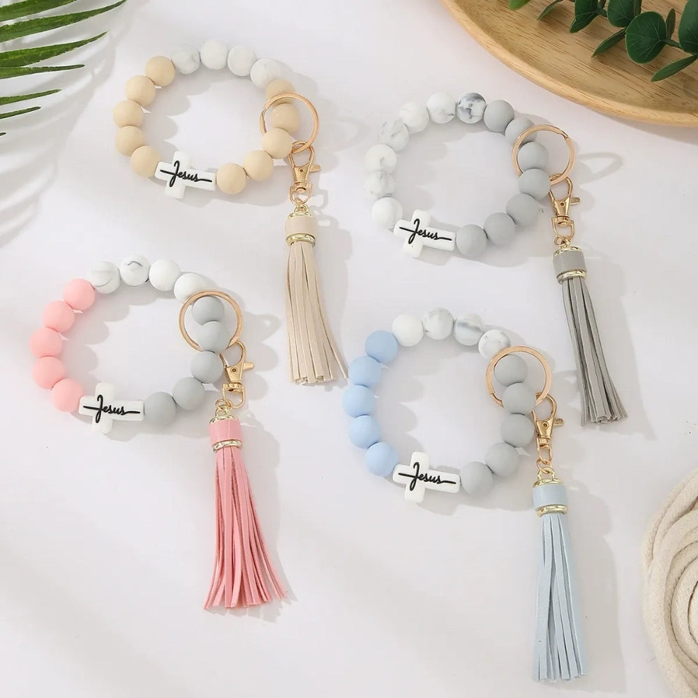 Four Jesus cross silicone bead bracelet keychains with leather tassels in beige, pink, blue, and gray marble color combinations on a white surface. - Random Mixed