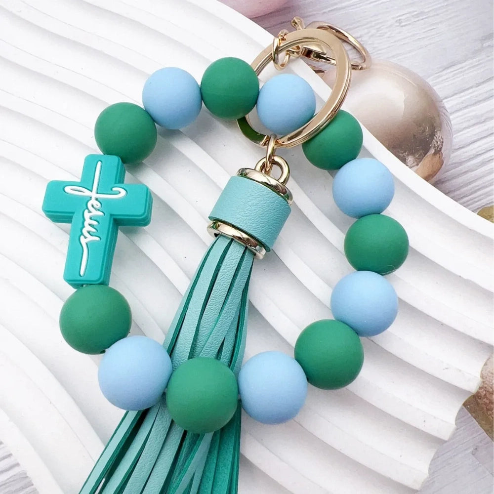 Colorful Jesus cross silicone bead stretch bracelets with tassel keychains in black, pink, purple, teal, and green color combinations on white background. - Green