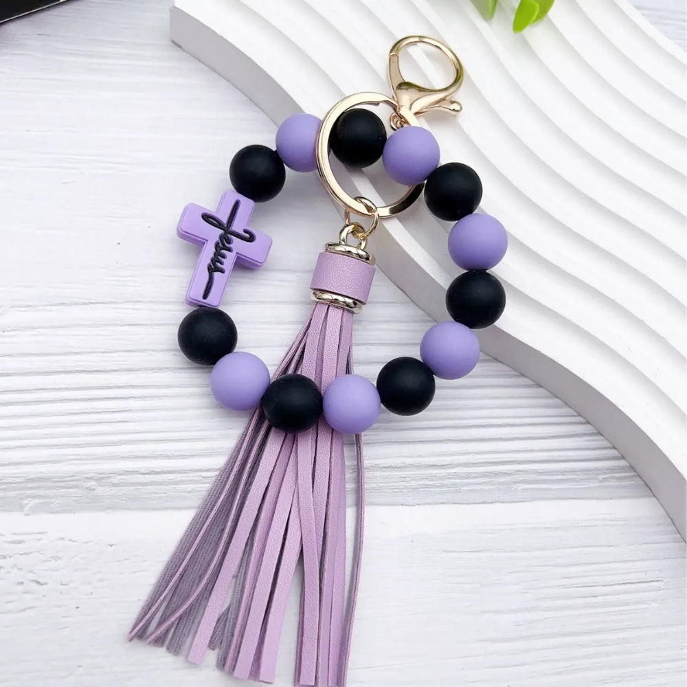 Colorful Jesus cross silicone bead stretch bracelets with tassel keychains in black, pink, purple, teal, and green color combinations on white background. - Purple