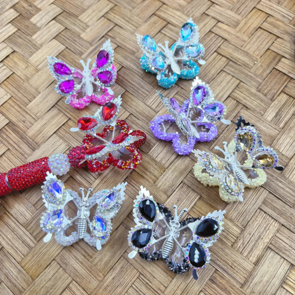 Large multicolor butterfly acrylic fancy beads with rhinestone wings in pink, blue, purple, red, white, cream, and black on a woven mat beside a red beadable pen.