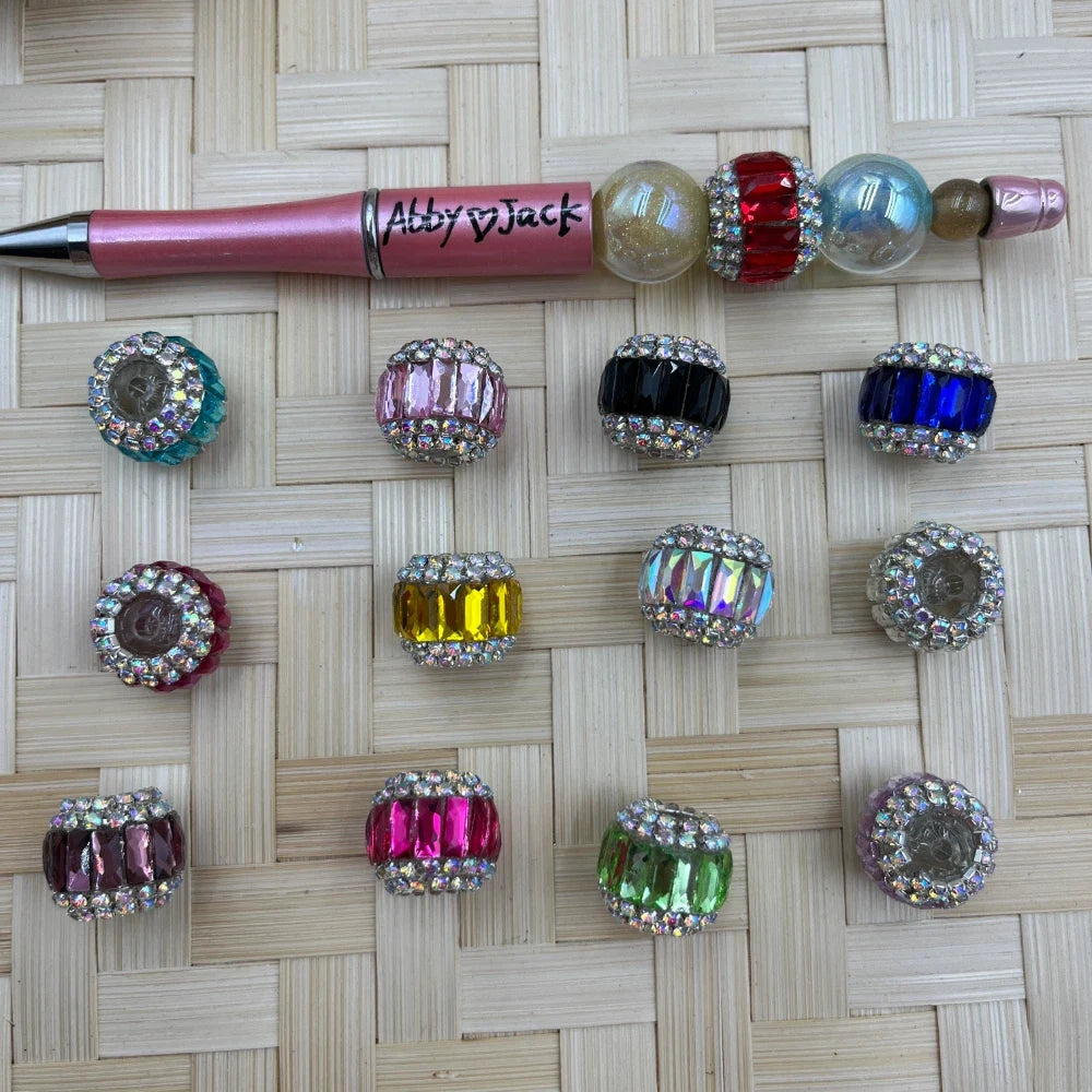 Assorted large hole faceted rhinestone acrylic barrel beads in pink, blue, black, green, yellow, purple and iridescent colors shown with a pink beadable pen.