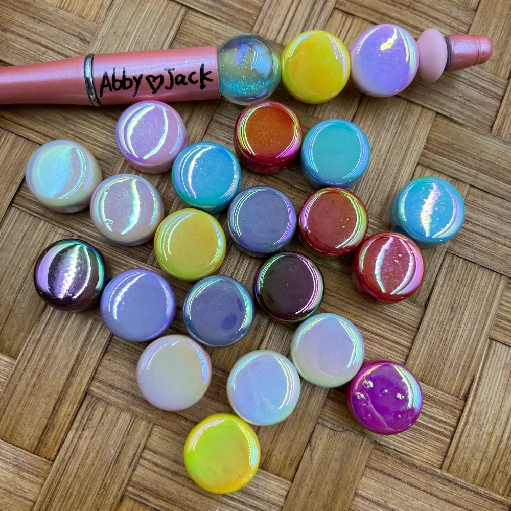 Assorted iridescent aurora shimmer acrylic beads in pastel and bold colors including purple, yellow, teal, red, lavender, and pink, displayed on a woven surface next to a beadable pen branded Abby Jack.