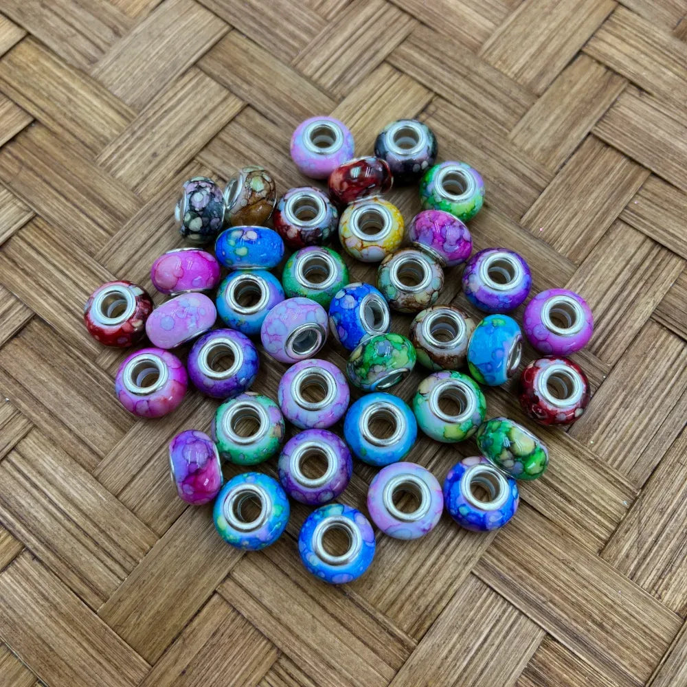Assorted colorful marble galaxy acrylic spacer beads with silver metal cores in pink, blue, purple, green, and multicolor patterns displayed on woven rattan surface
