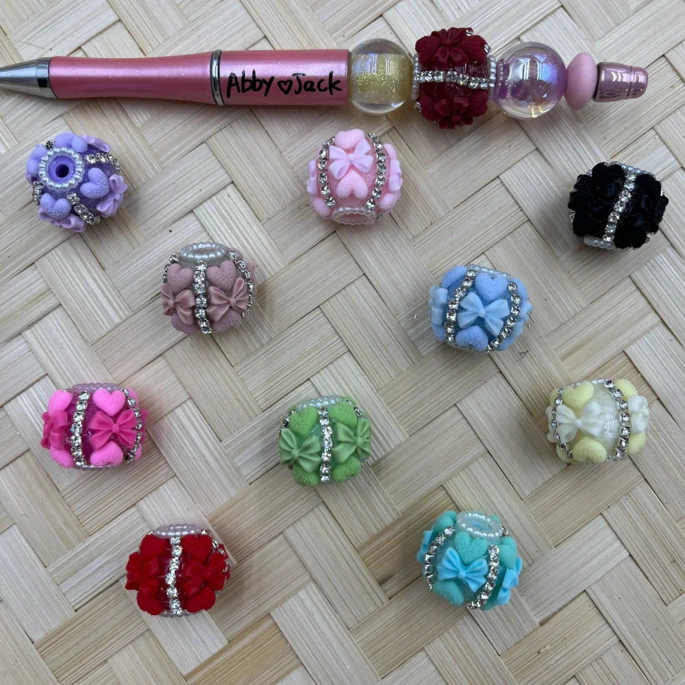 Assorted colorful clay fancy beads decorated with rhinestone bands, bow and heart embellishments in purple, pink, mauve, hot pink, red, green, blue, teal, yellow, and black, displayed next to a beadable pen on a wicker surface.