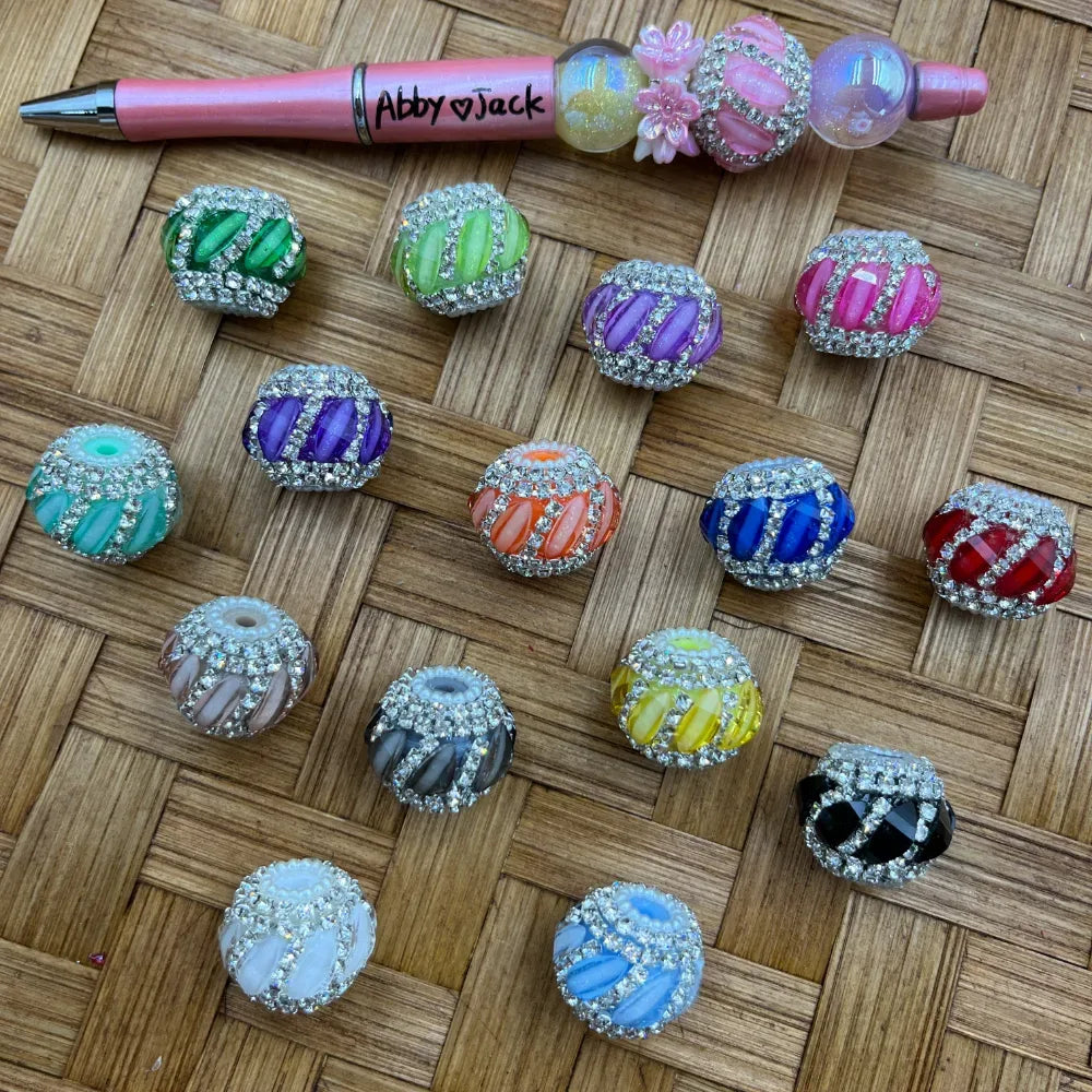 Assorted rhinestone encrusted striped acrylic round beads in 14 colors including green, purple, orange, blue, yellow, red, black, white, and pink, displayed on a woven surface next to a decorated beadable pen.