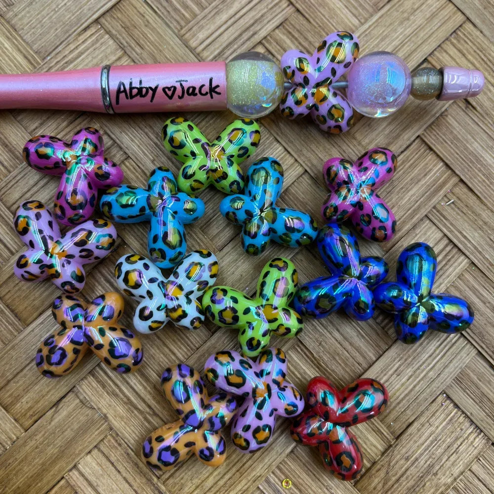 Colorful leopard print butterfly acrylic beads in multiple colors including pink, blue, green, purple, orange, and red displayed on a woven surface next to a beadable pen.