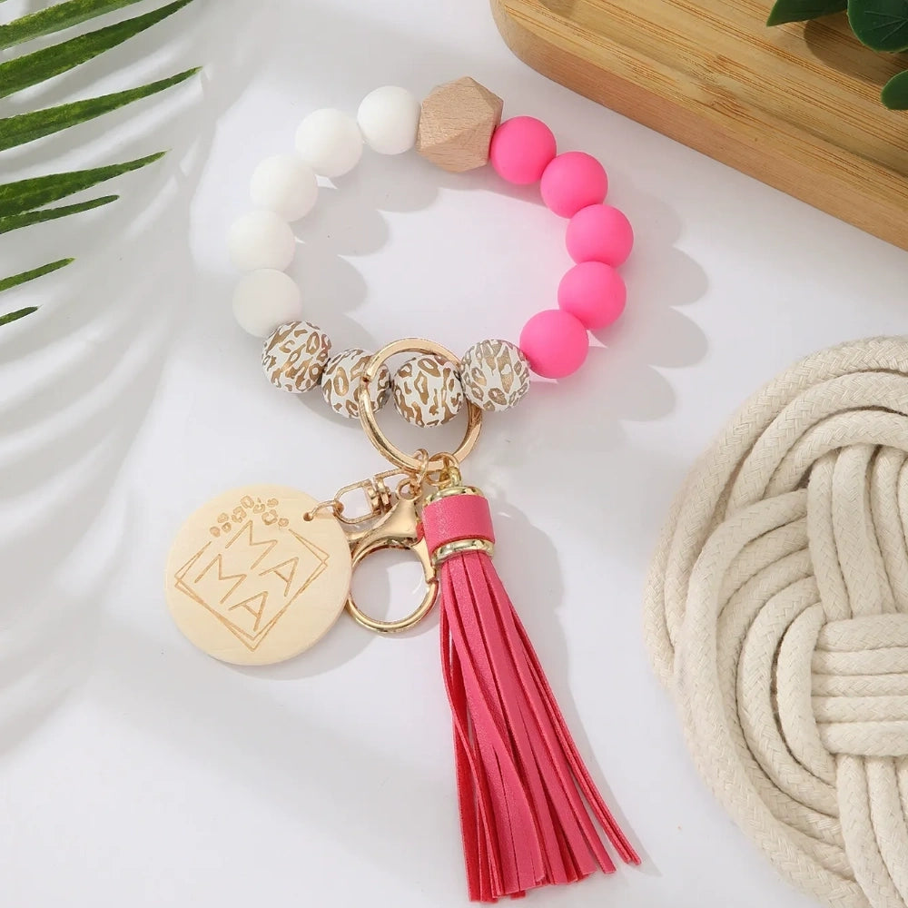 Assorted colorful silicone bead wristlet keychains featuring leopard print beads, leather tassels, and engraved gold Mama charm pendants in multiple color combinations including orange, purple, pink, blue, black, mint, and gray arranged on a white surface. - A