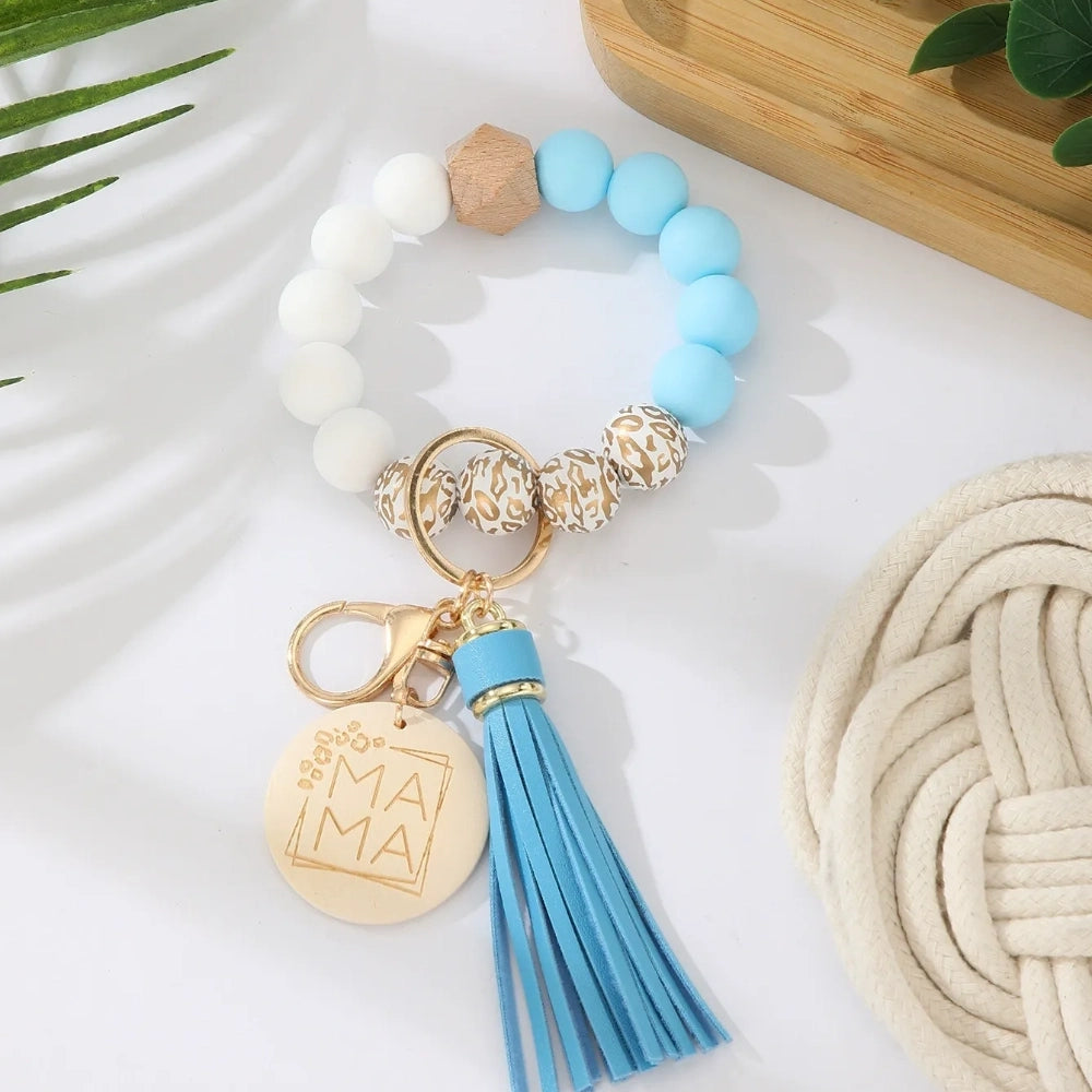 Assorted colorful silicone bead wristlet keychains featuring leopard print beads, leather tassels, and engraved gold Mama charm pendants in multiple color combinations including orange, purple, pink, blue, black, mint, and gray arranged on a white surface. - B