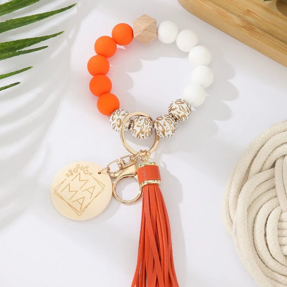 Assorted colorful silicone bead wristlet keychains featuring leopard print beads, leather tassels, and engraved gold Mama charm pendants in multiple color combinations including orange, purple, pink, blue, black, mint, and gray arranged on a white surface. - C