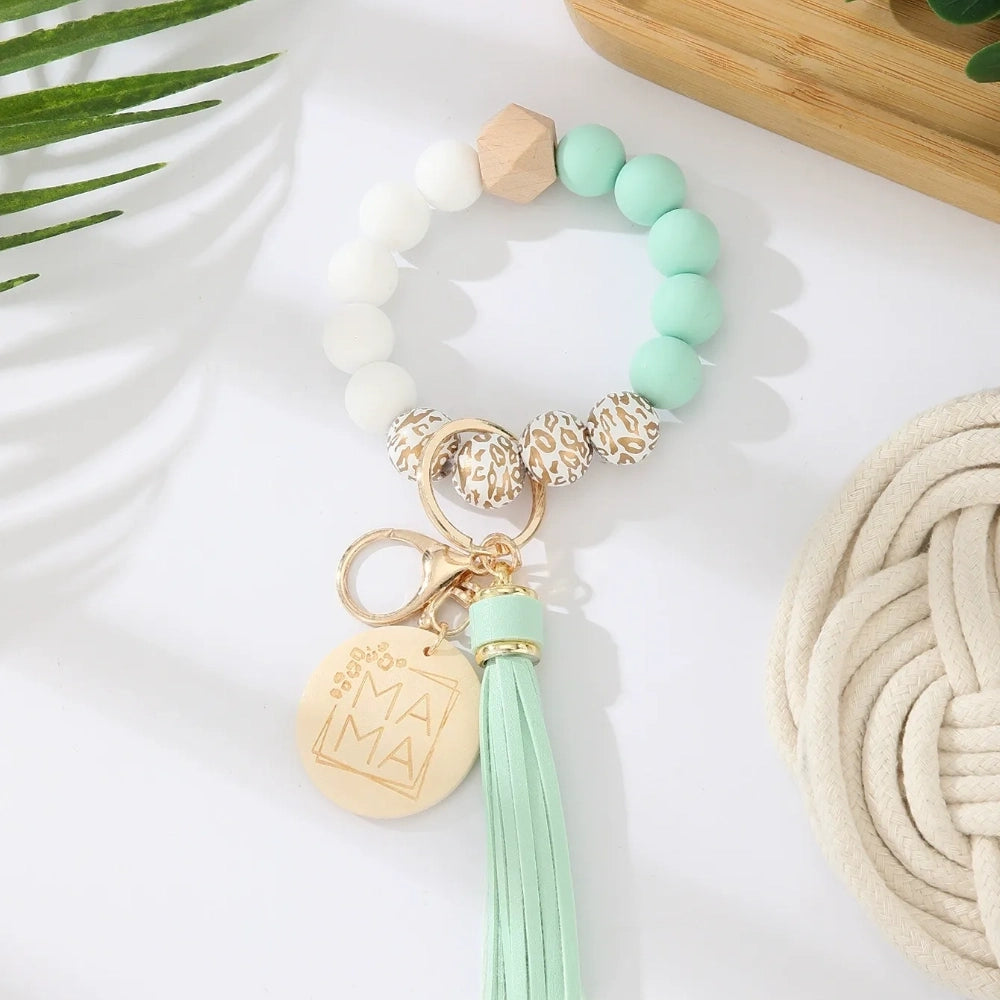 Assorted colorful silicone bead wristlet keychains featuring leopard print beads, leather tassels, and engraved gold Mama charm pendants in multiple color combinations including orange, purple, pink, blue, black, mint, and gray arranged on a white surface. - D