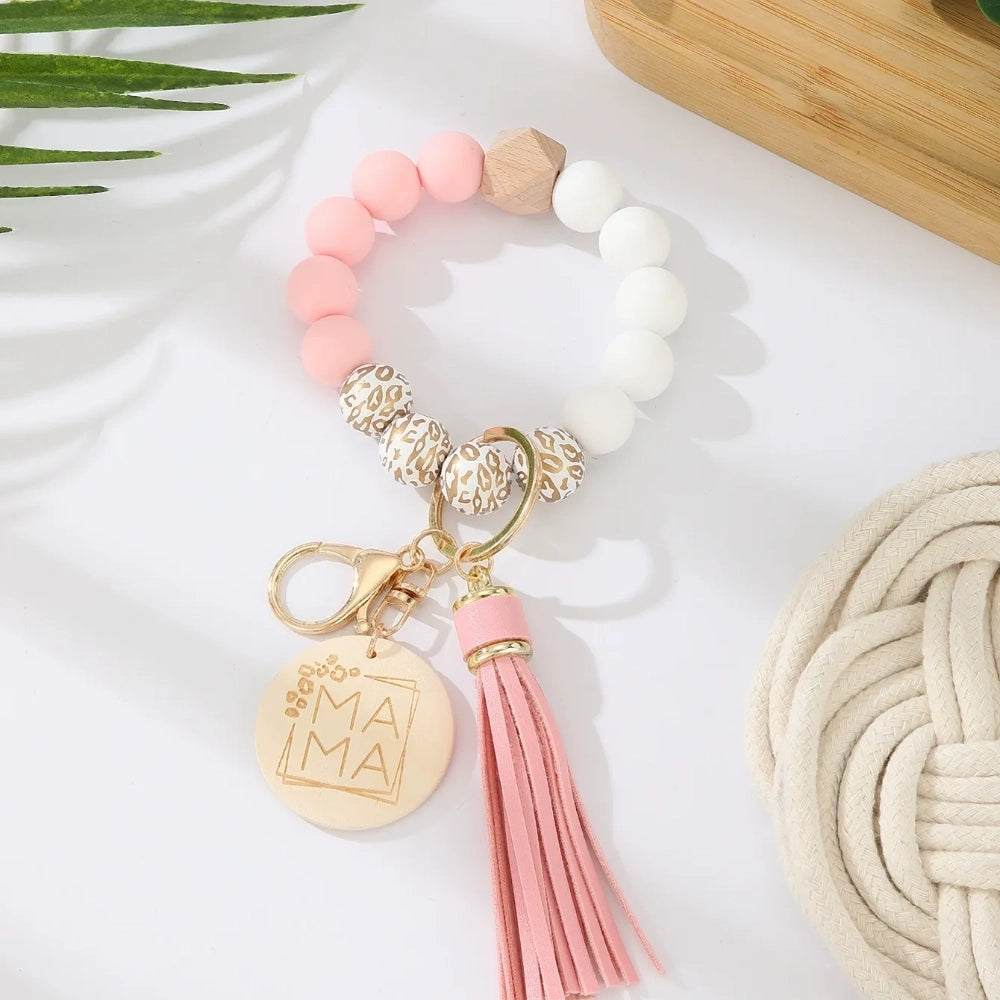 Assorted colorful silicone bead wristlet keychains featuring leopard print beads, leather tassels, and engraved gold Mama charm pendants in multiple color combinations including orange, purple, pink, blue, black, mint, and gray arranged on a white surface. - E