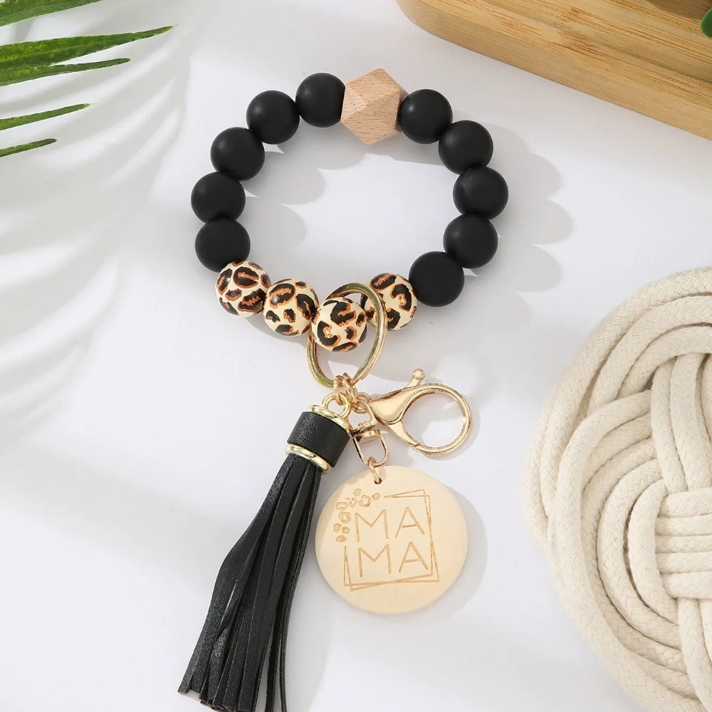 Assorted colorful silicone bead wristlet keychains featuring leopard print beads, leather tassels, and engraved gold Mama charm pendants in multiple color combinations including orange, purple, pink, blue, black, mint, and gray arranged on a white surface. - F