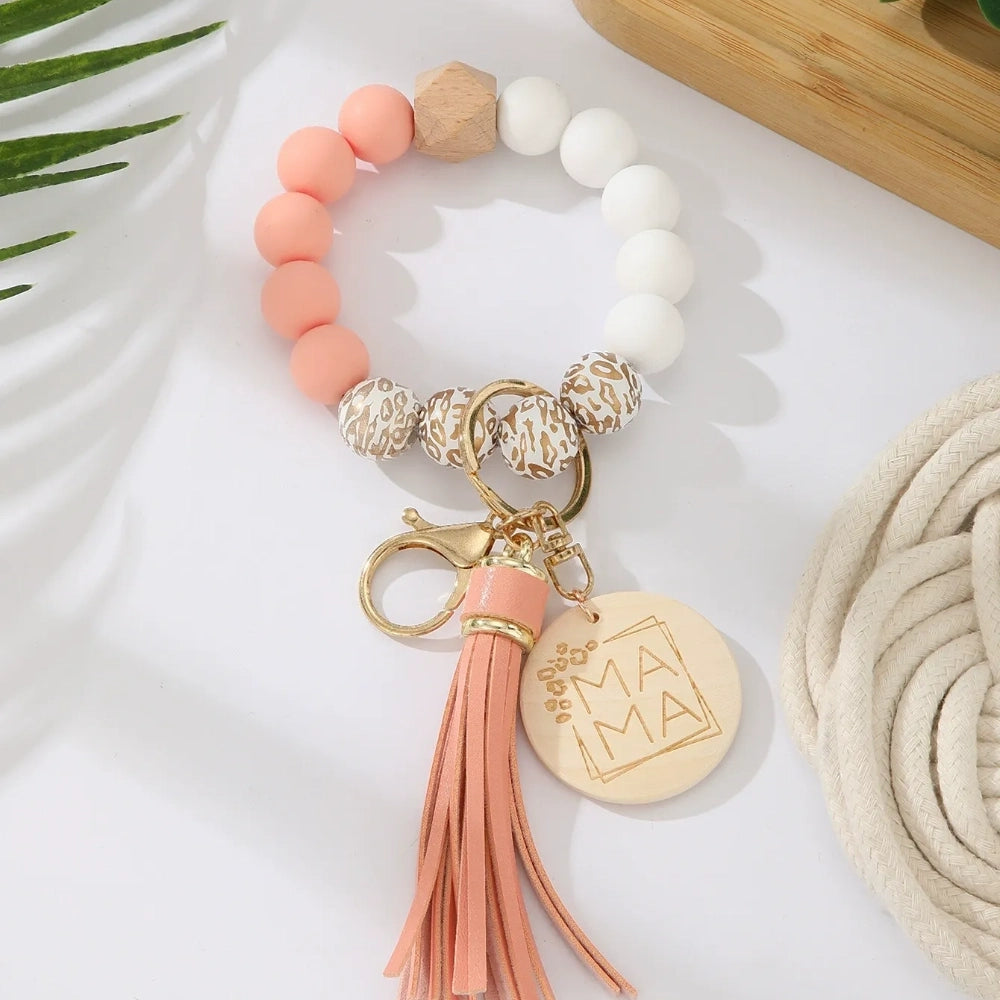 Assorted colorful silicone bead wristlet keychains featuring leopard print beads, leather tassels, and engraved gold Mama charm pendants in multiple color combinations including orange, purple, pink, blue, black, mint, and gray arranged on a white surface. - G