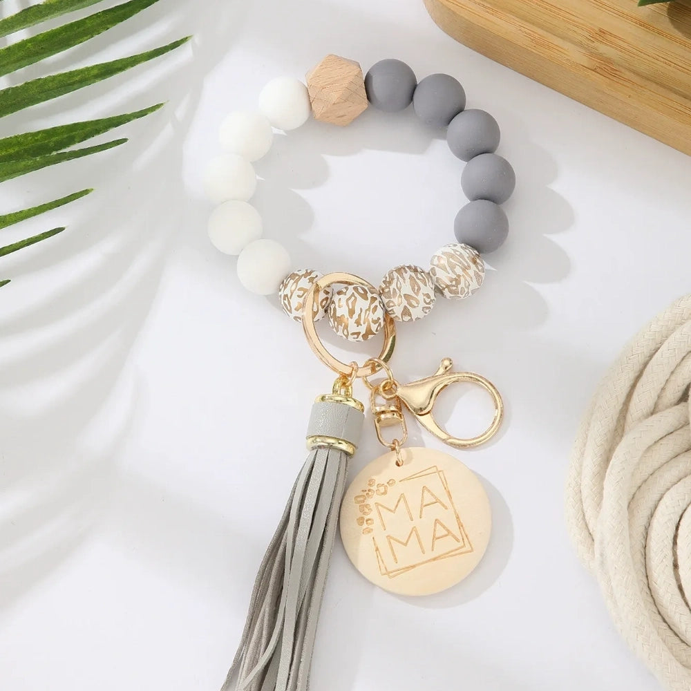 Assorted colorful silicone bead wristlet keychains featuring leopard print beads, leather tassels, and engraved gold Mama charm pendants in multiple color combinations including orange, purple, pink, blue, black, mint, and gray arranged on a white surface. - K