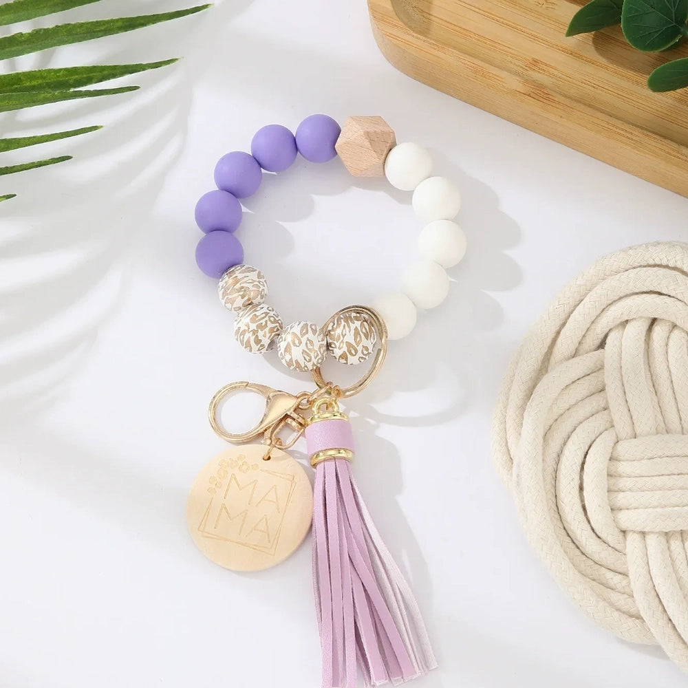 Assorted colorful silicone bead wristlet keychains featuring leopard print beads, leather tassels, and engraved gold Mama charm pendants in multiple color combinations including orange, purple, pink, blue, black, mint, and gray arranged on a white surface. - L