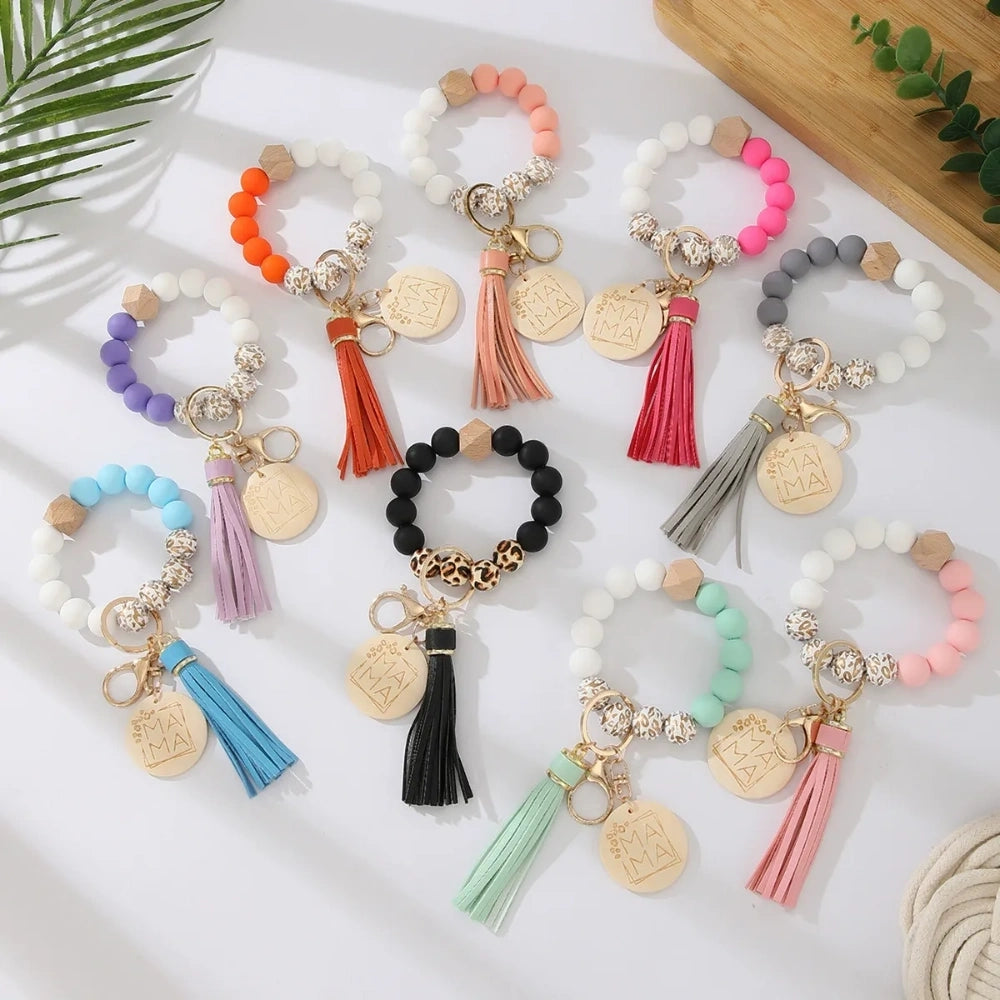 Assorted colorful silicone bead wristlet keychains featuring leopard print beads, leather tassels, and engraved gold Mama charm pendants in multiple color combinations including orange, purple, pink, blue, black, mint, and gray arranged on a white surface. - Random Mixed