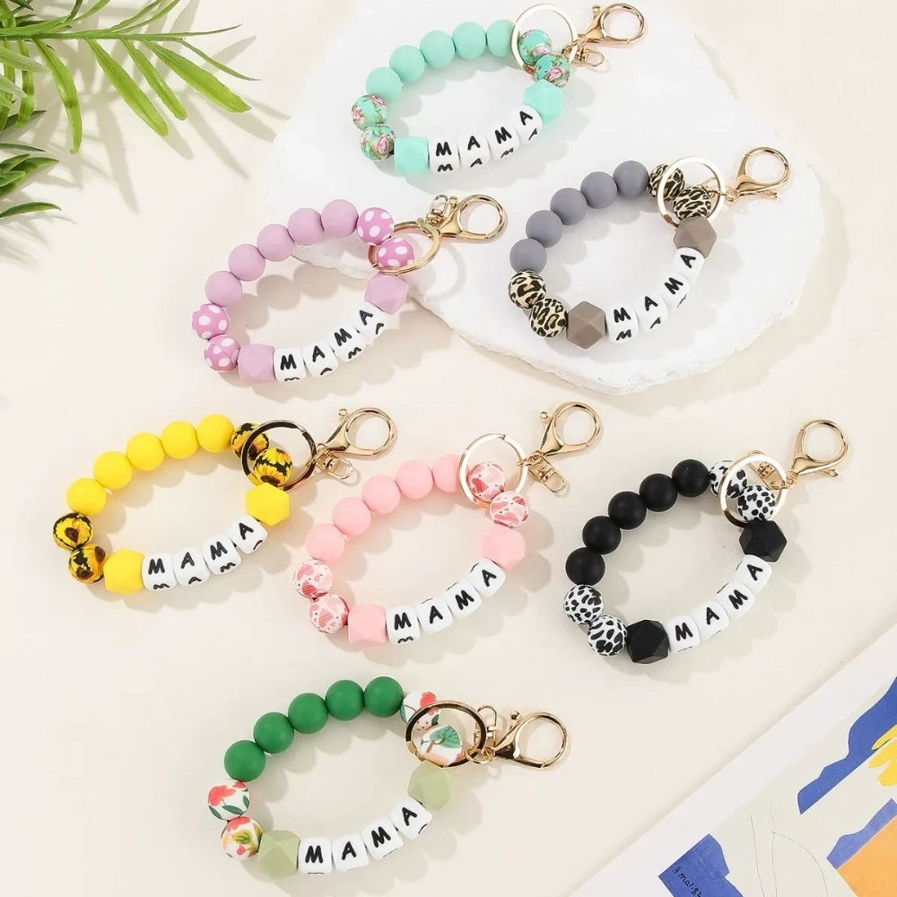 Seven MAMA letter silicone bead wristlet keychains in assorted colors including mint, lavender, gray, yellow, pink, black, and green with patterned accent beads and gold lobster clasp hardware on a light background.