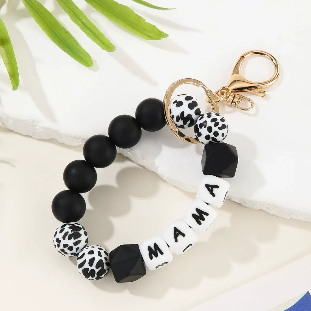 Seven MAMA letter silicone bead wristlet keychains in assorted colors including mint, lavender, gray, yellow, pink, black, and green with patterned accent beads and gold lobster clasp hardware on a light background. - Black