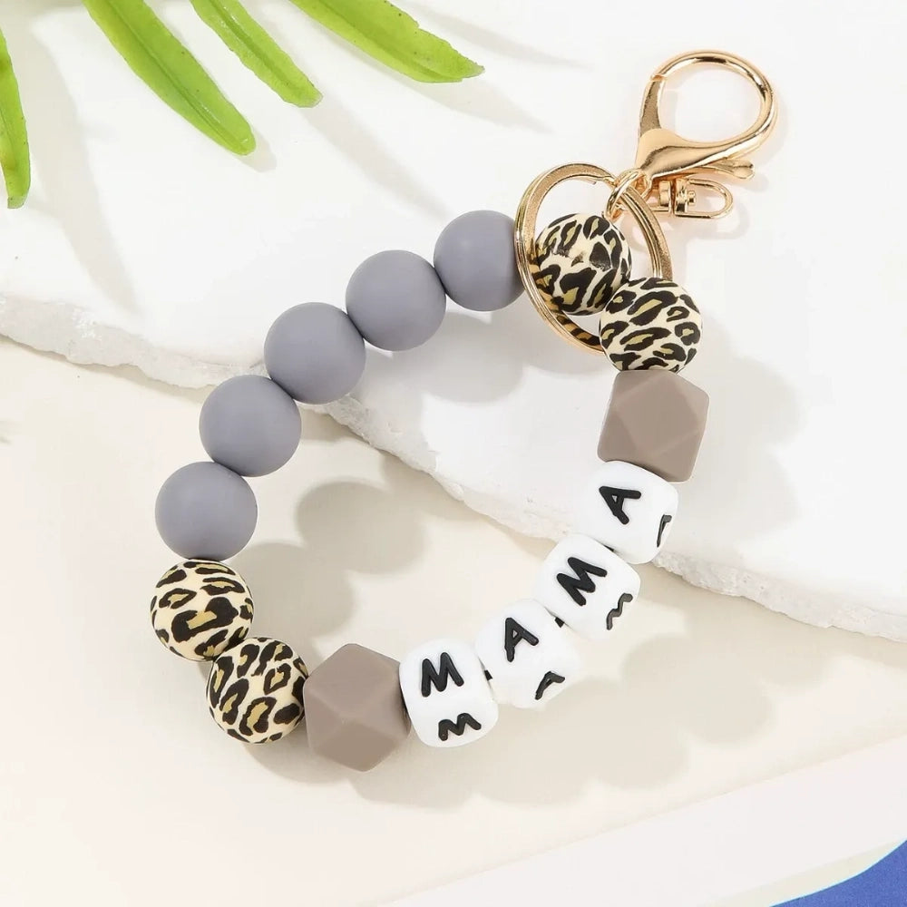 Seven MAMA letter silicone bead wristlet keychains in assorted colors including mint, lavender, gray, yellow, pink, black, and green with patterned accent beads and gold lobster clasp hardware on a light background. - Gray