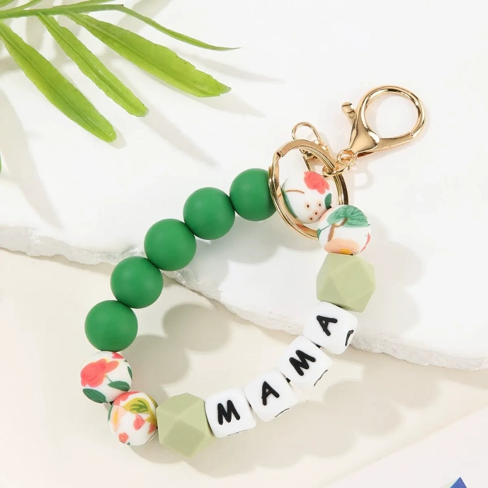 Seven MAMA letter silicone bead wristlet keychains in assorted colors including mint, lavender, gray, yellow, pink, black, and green with patterned accent beads and gold lobster clasp hardware on a light background. - Green