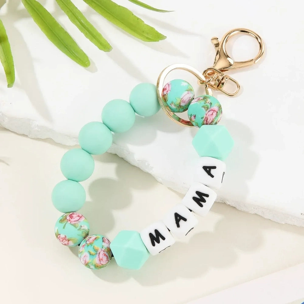 Seven MAMA letter silicone bead wristlet keychains in assorted colors including mint, lavender, gray, yellow, pink, black, and green with patterned accent beads and gold lobster clasp hardware on a light background. - Light Green
