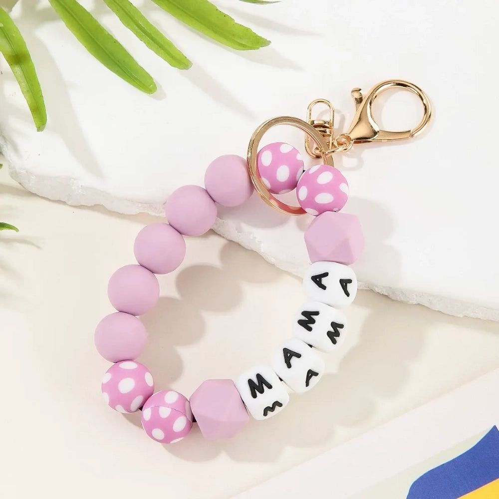 Seven MAMA letter silicone bead wristlet keychains in assorted colors including mint, lavender, gray, yellow, pink, black, and green with patterned accent beads and gold lobster clasp hardware on a light background. - Purple